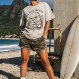 a woman standing in front of a life guard tower wearing a faded green graphic tee with a monk seal design