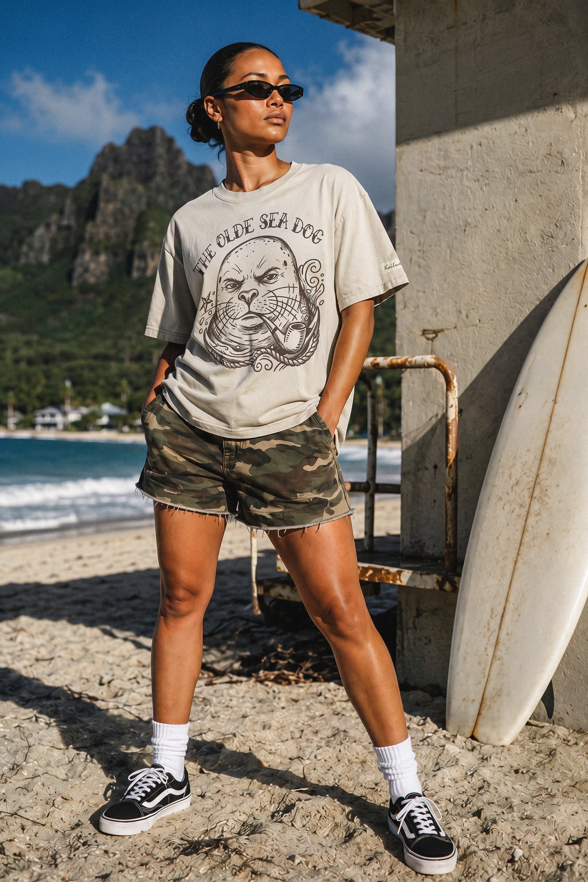 a woman standing in front of a life guard tower wearing a faded green graphic tee with a monk seal design