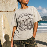 woman standing in front of a life guard tower on the beach wearing a faded green graphic tee with a monk seal design