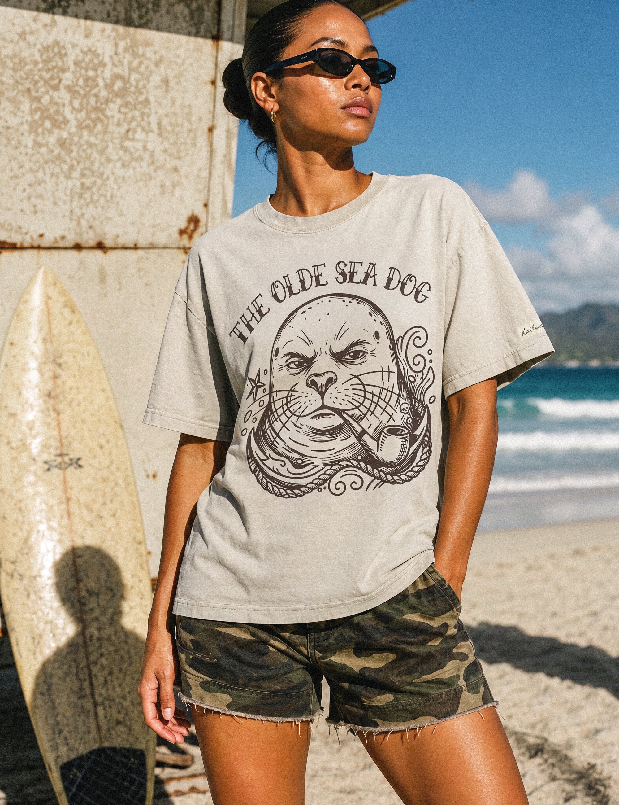 woman standing in front of a life guard tower on the beach wearing a faded green graphic tee with a monk seal design
