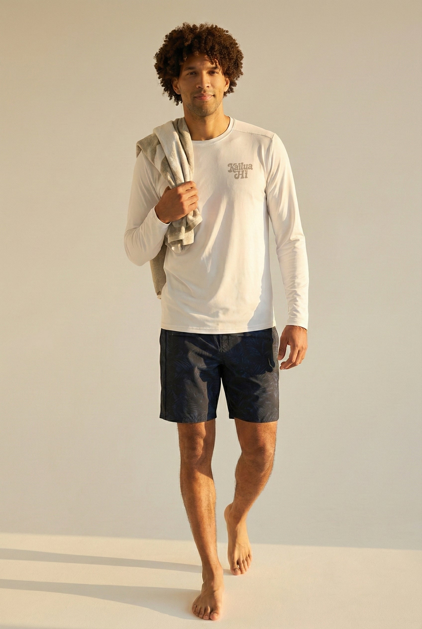 man wearing white long sleeve shirt with gray kailua hi logo on chest and black shorts with a towel over his shoulder
