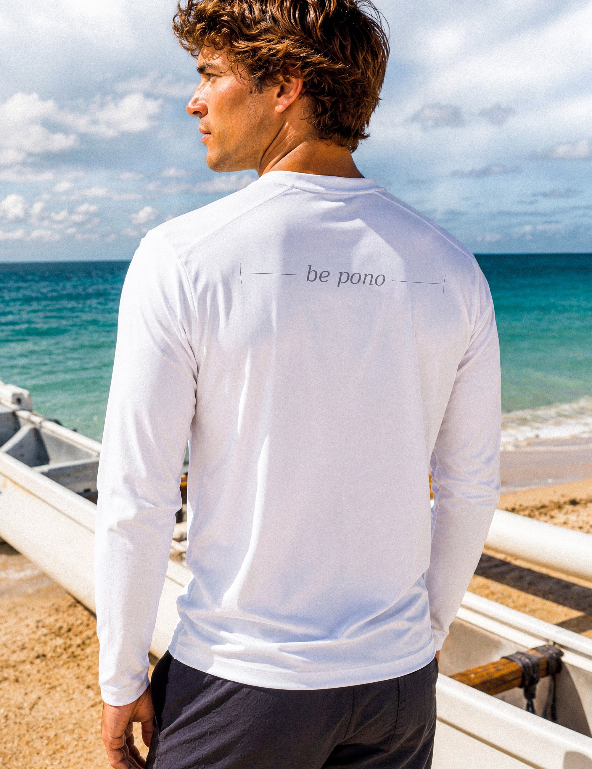 A man wearing a white long-sleeve shirt with 'be pono' graphic on the beach.