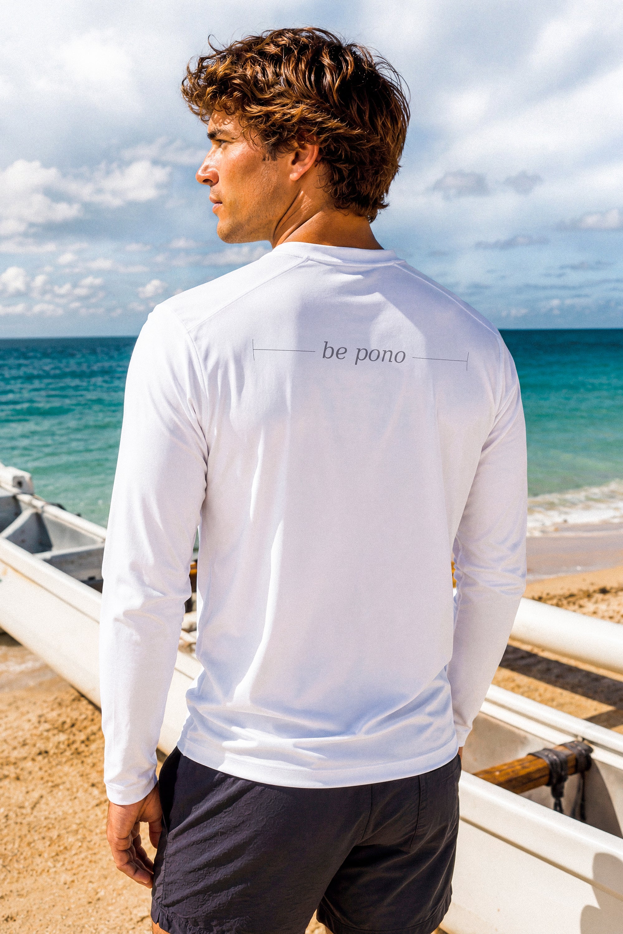 A man wearing a white long-sleeve shirt with 'be pono' graphic on the beach.