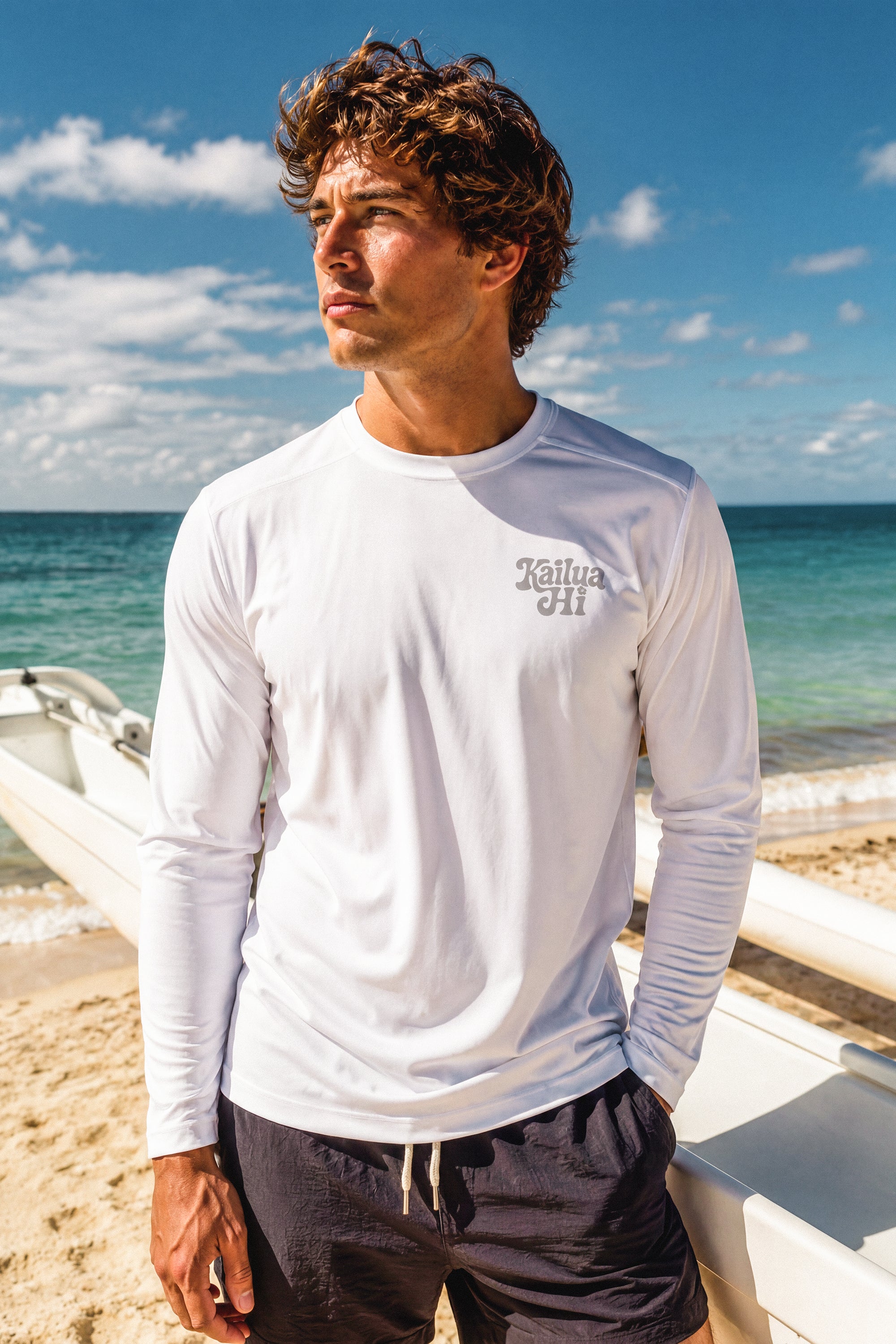 Man wearing a white long-sleeve shirt with a Kailua Hi logo on the left chest on a beach