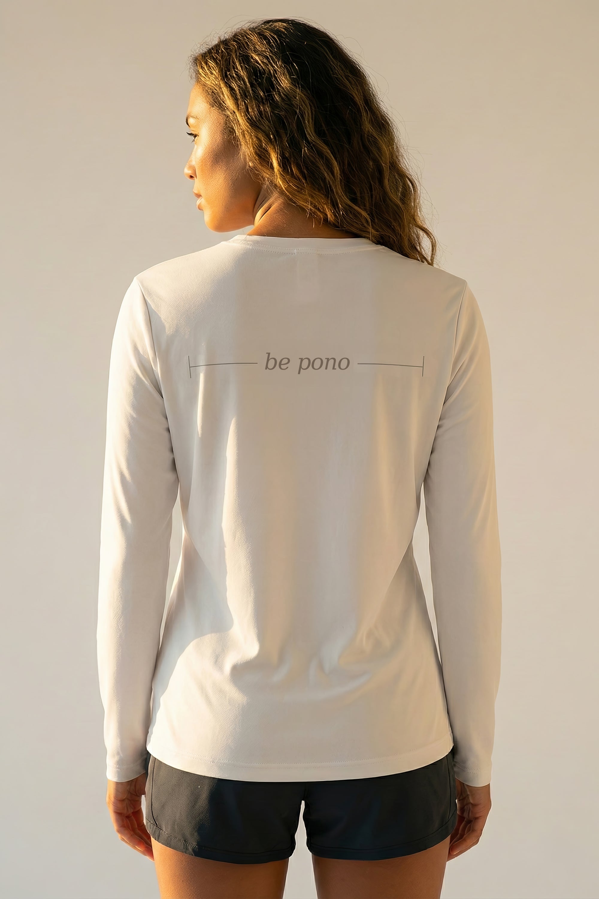 woman wearing a white long-sleeve shirt with grey 'be pono' text on the back against a plain background
