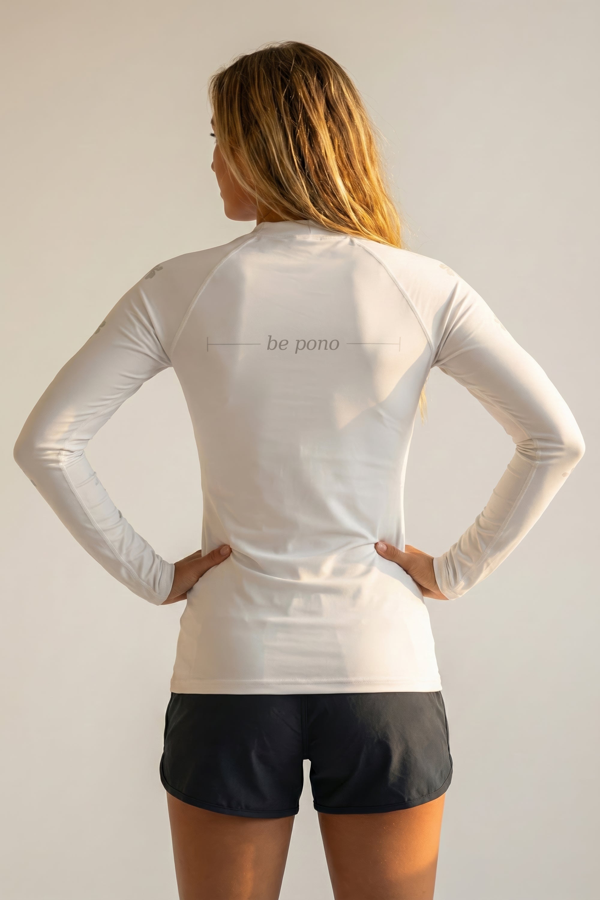 Person wearing a white long-sleeve athletic shirt with grey 'be pono' text on the back, standing against a plain background.