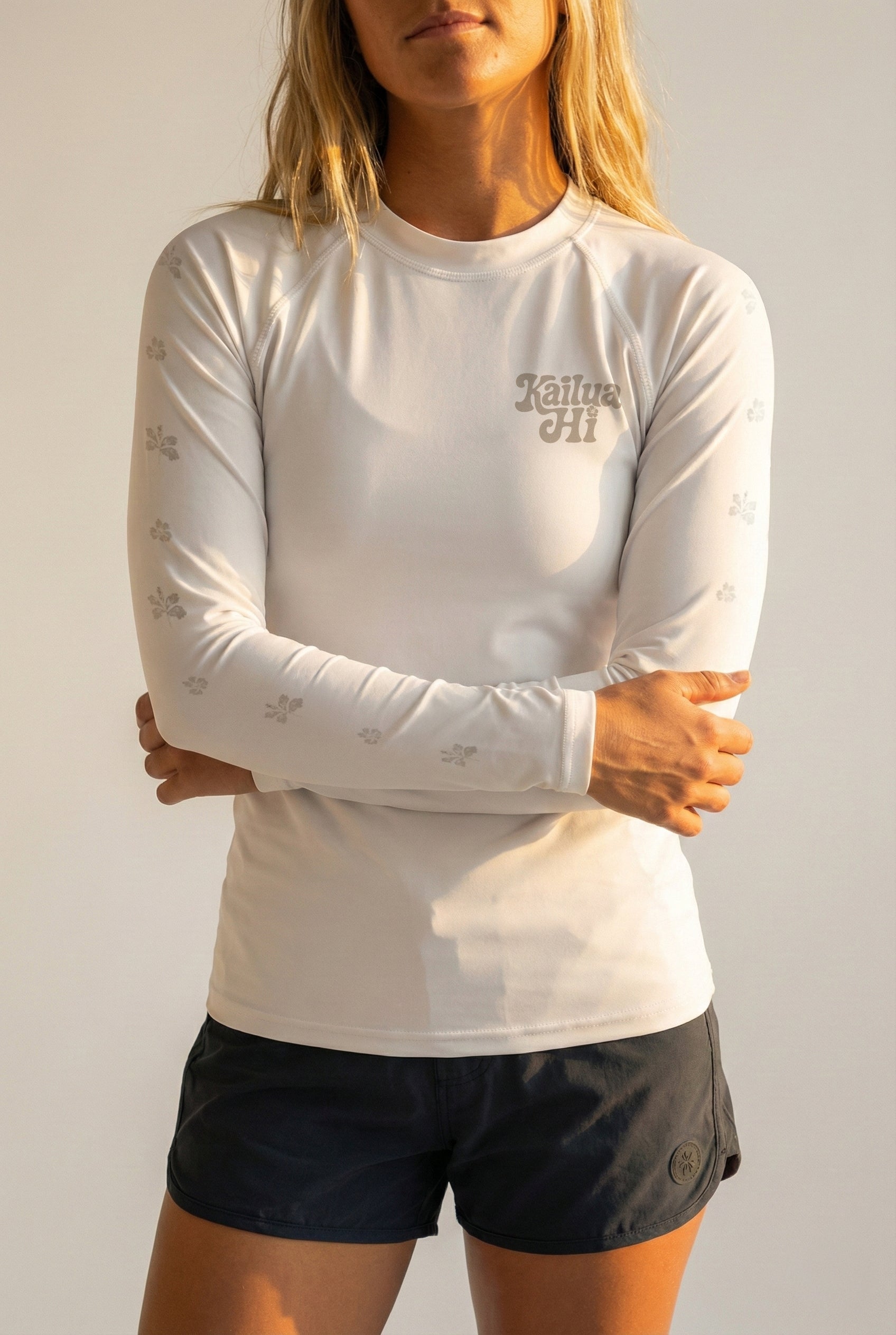 woman wearing a white long-sleeve shirt with a Kailua Hi logo and black shorts against a plain background