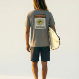 man holding a surfboard wearing a grey short sleeve tee with a warning sign graphic design on back