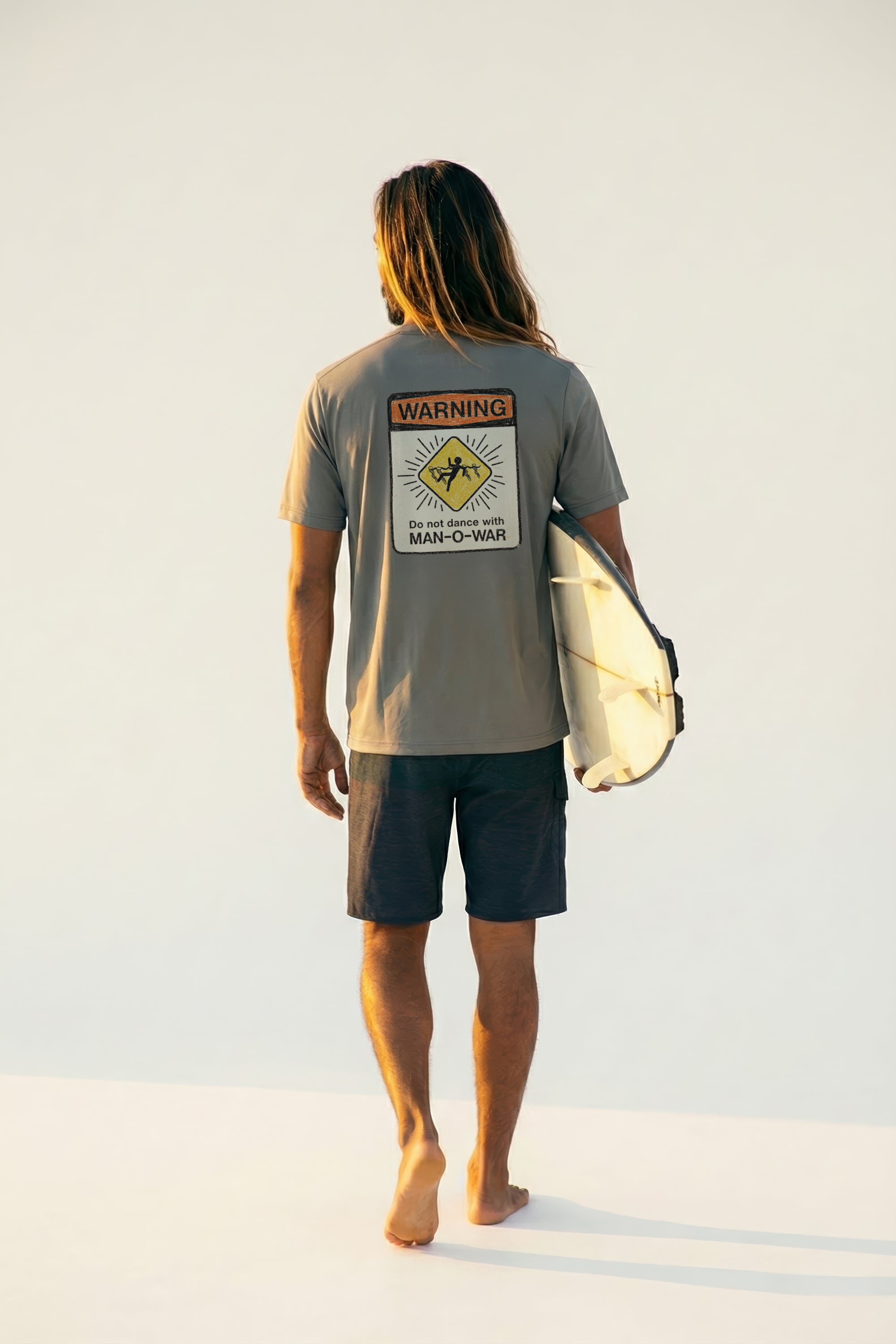 man holding a surfboard wearing a grey short sleeve tee with a warning sign graphic design on back
