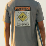 man holding a surfboard wearing a grey short sleeve tee with a warning sign graphic design on back