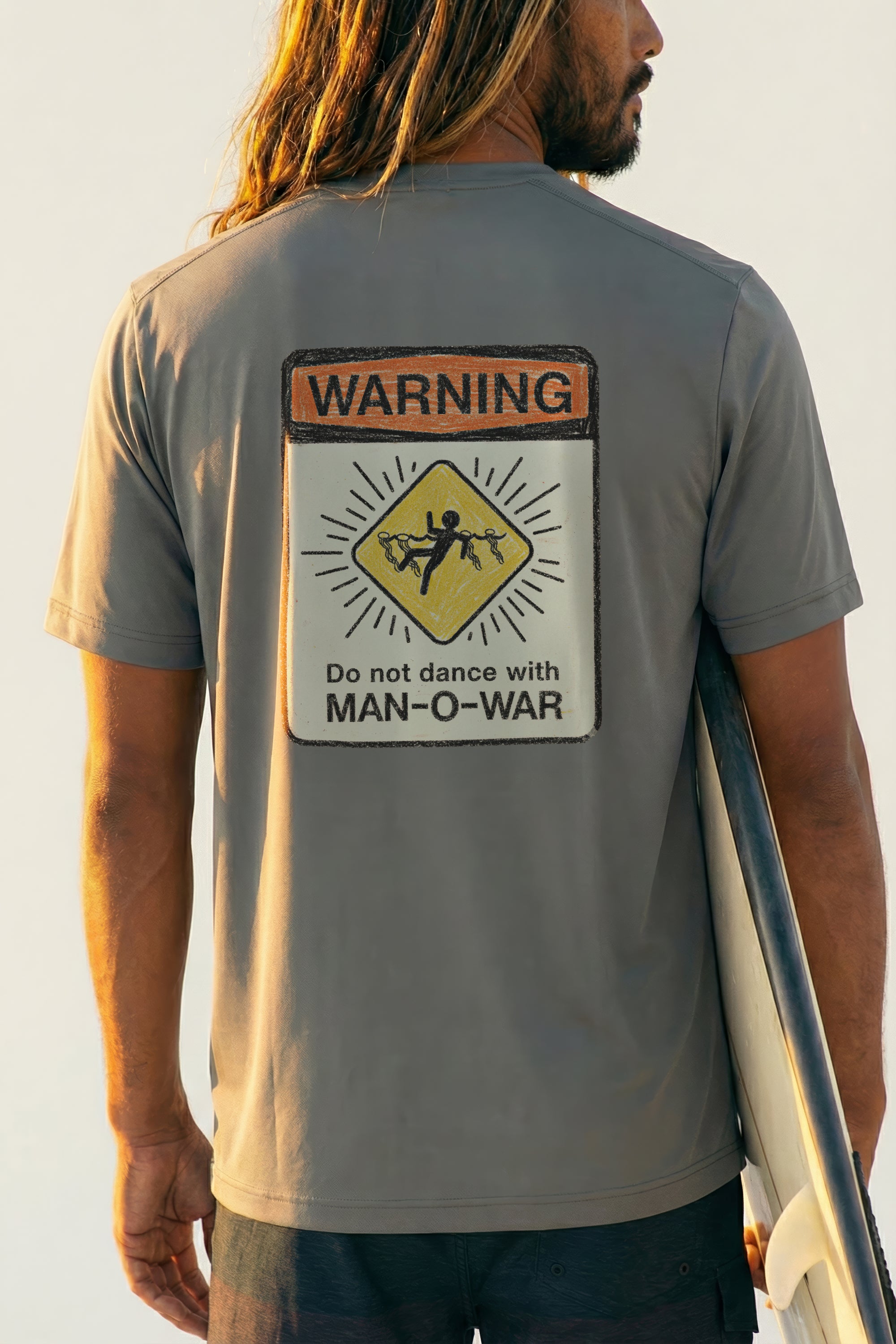 man holding a surfboard wearing a grey short sleeve tee with a warning sign graphic design on back