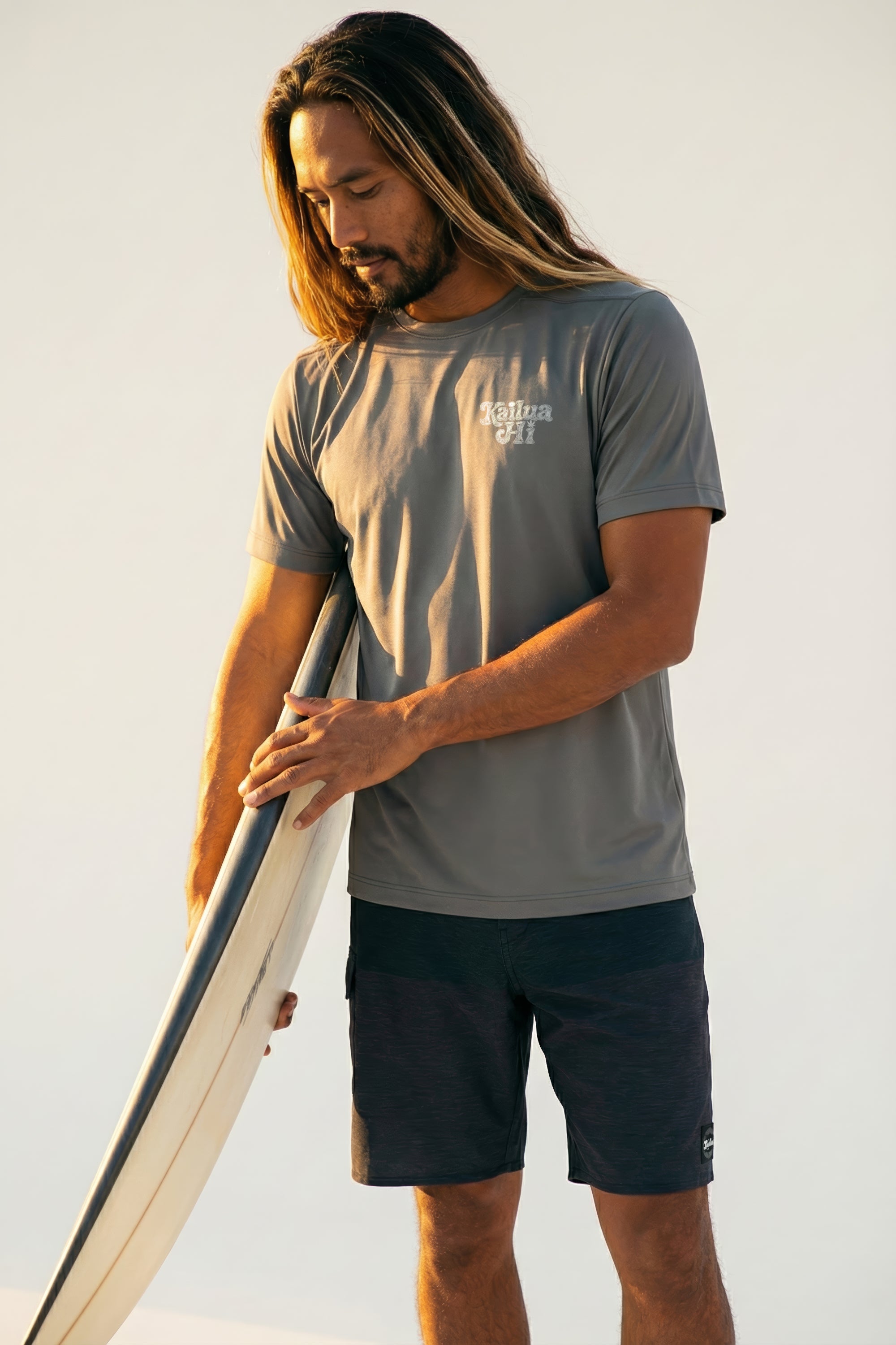man holding a surfboard wearing a grey short sleeve tee with a kailua hi logo on front