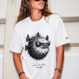 a woman wearing a white shirt with a puffer fish graphic standing in front of a surfboard 
