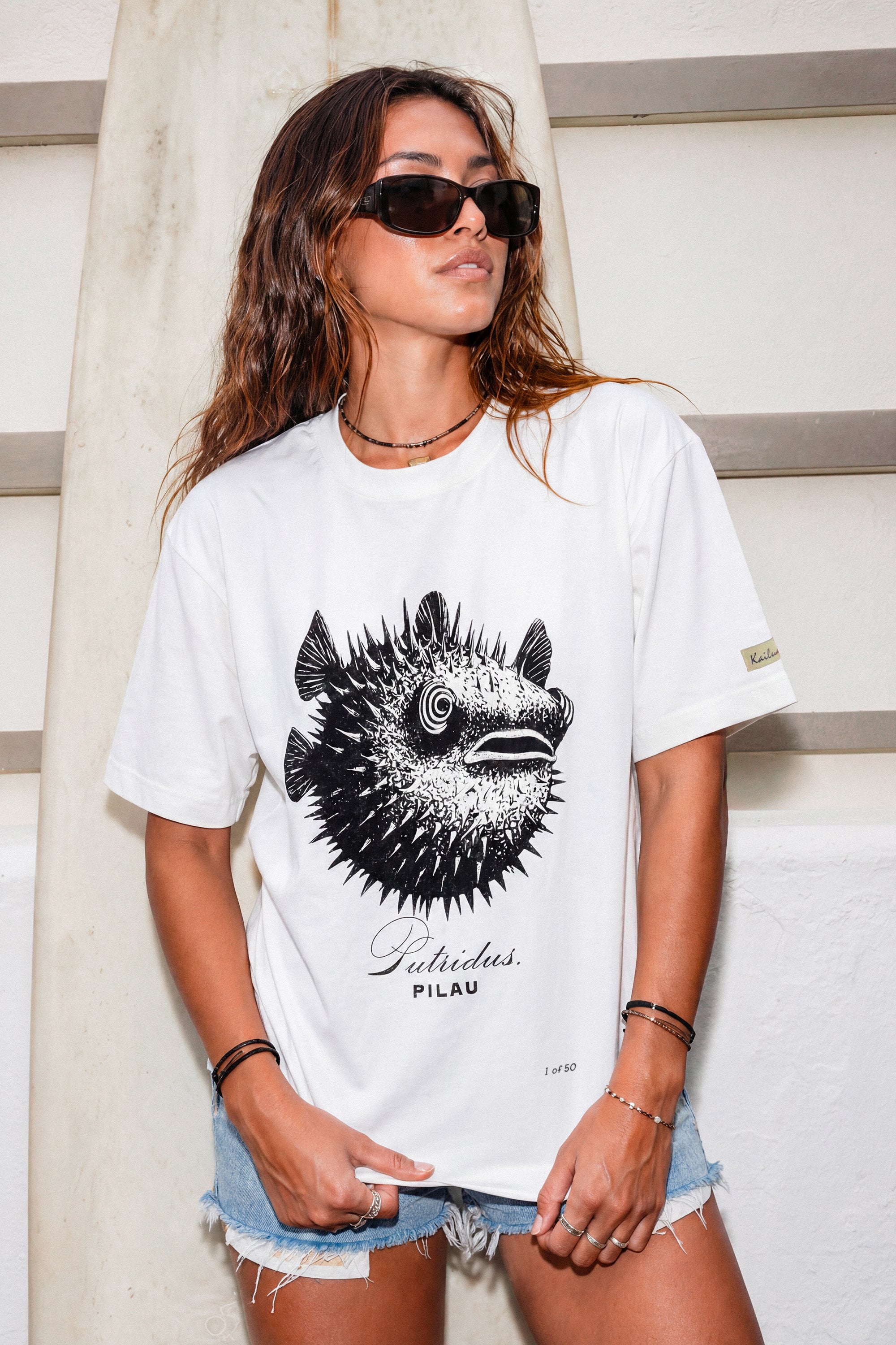 a woman wearing a white shirt with a puffer fish graphic standing in front of a surfboard 