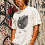 a man wearing a bone colored t-shirt with a puffer fish graphic standing against a wall covered with graffiti