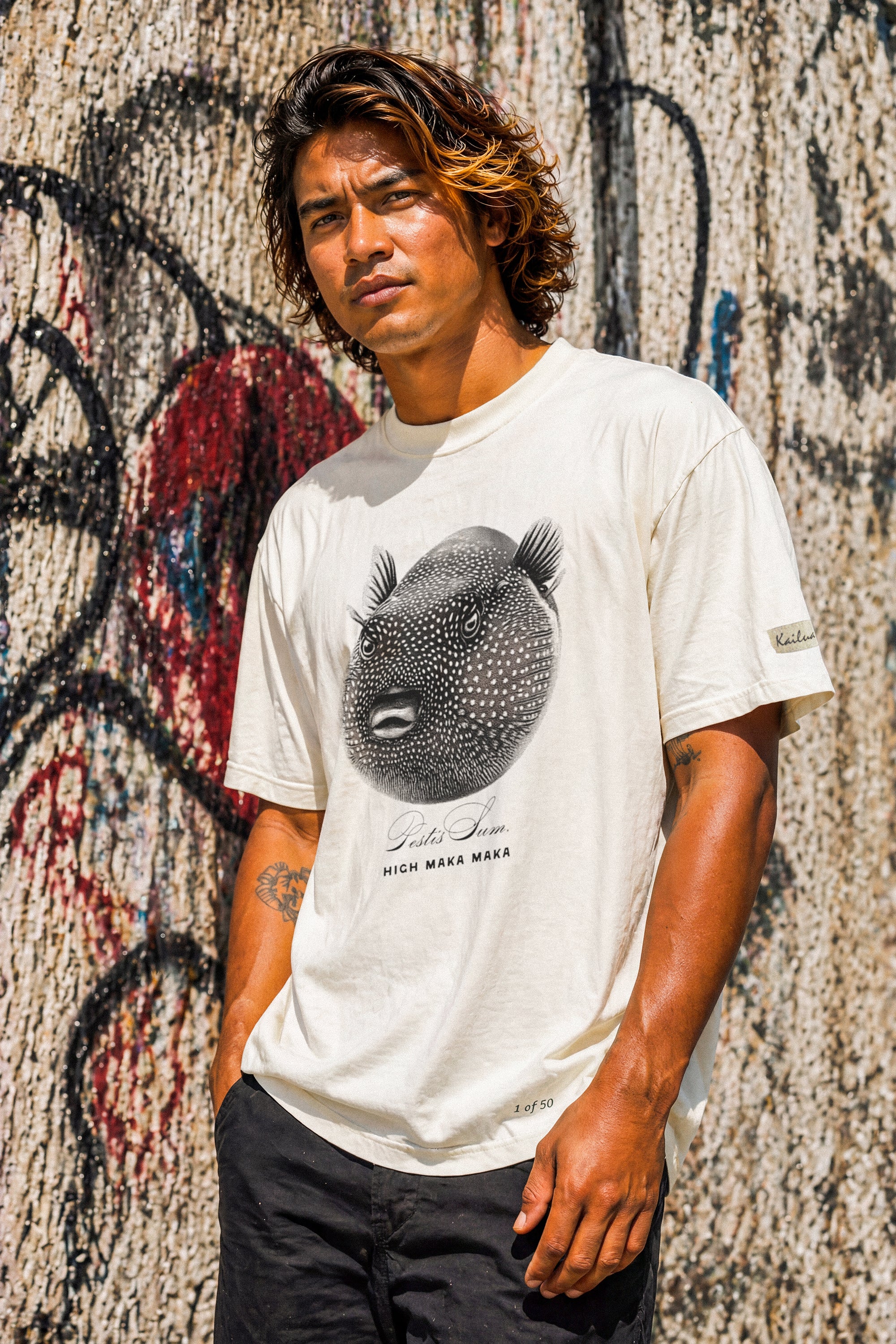 a man wearing a bone colored t-shirt with a puffer fish graphic standing against a wall covered with graffiti