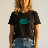 Woman wearing a black t-shirt with a blue fish graphic and denim shorts against a plain background