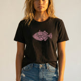 Woman wearing a black t-shirt with a pink fish design and denim shorts against a plain background