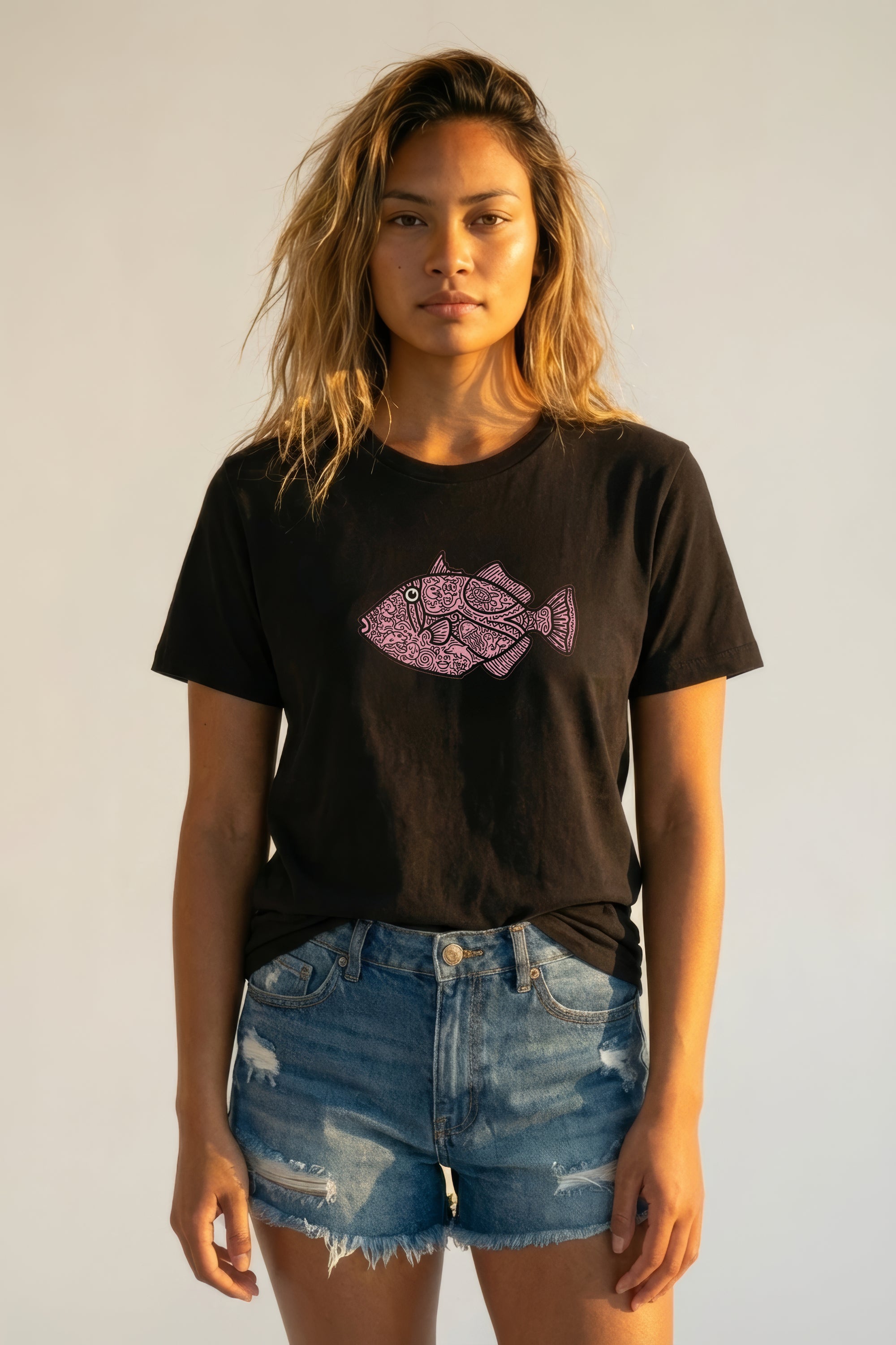 Woman wearing a black t-shirt with a pink fish design and denim shorts against a plain background