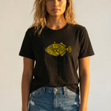 Woman wearing a black t-shirt with a yellow fish graphic and denim shorts against a plain background