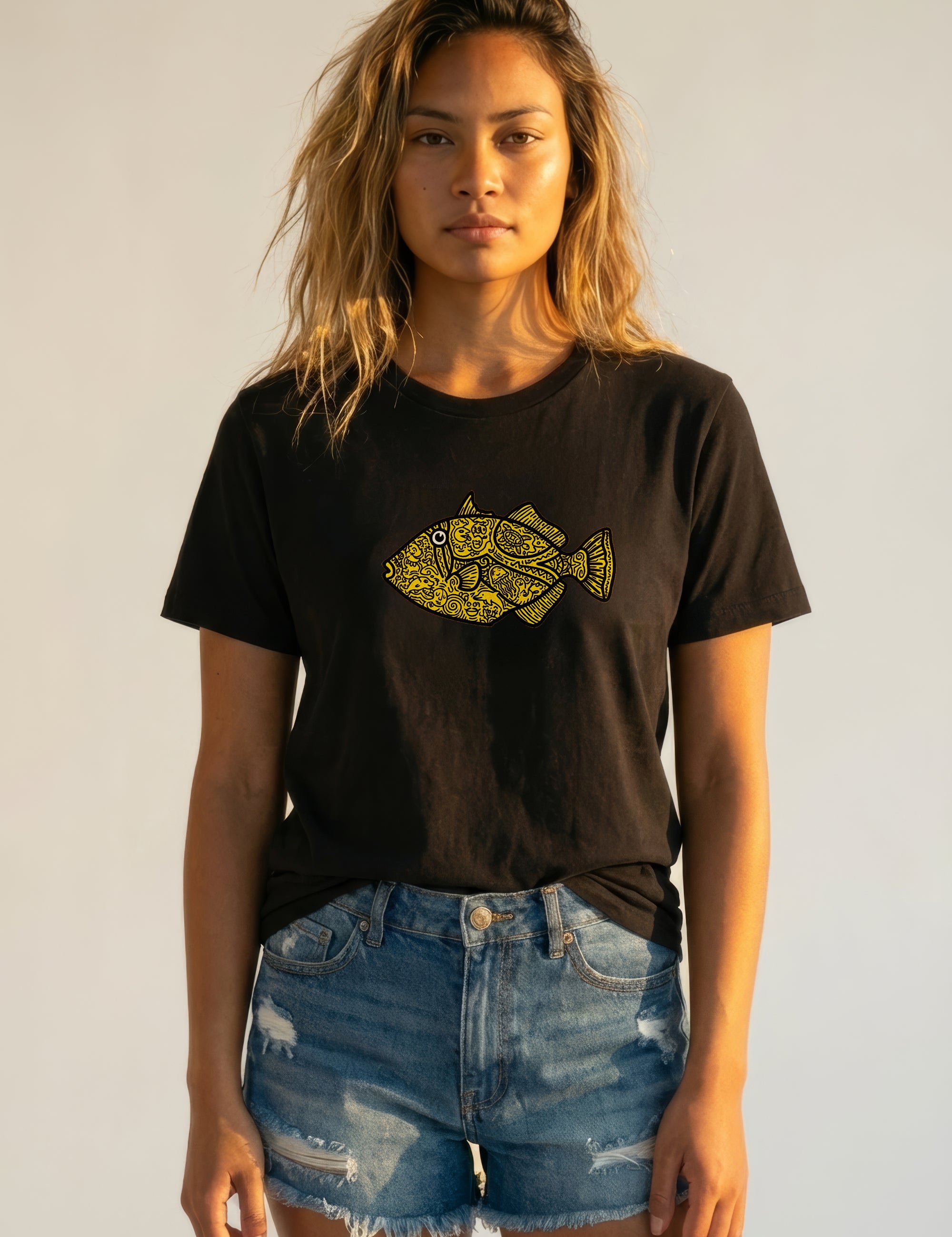 Woman wearing a black t-shirt with a yellow fish graphic and denim shorts against a plain background