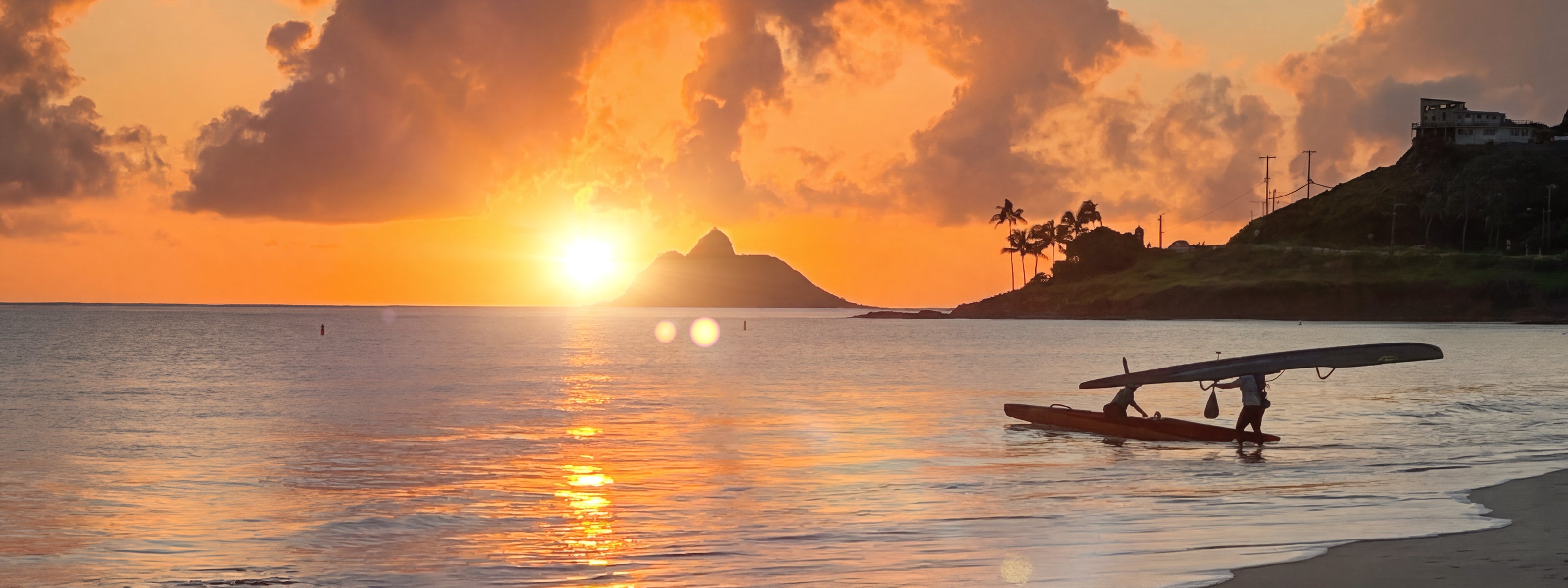 Sunset over a beach with a person in a canoe and palm trees in the background.