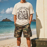 a man standing on the beach next to a surfboard wearing a faded green graphic tee with a monk seal design