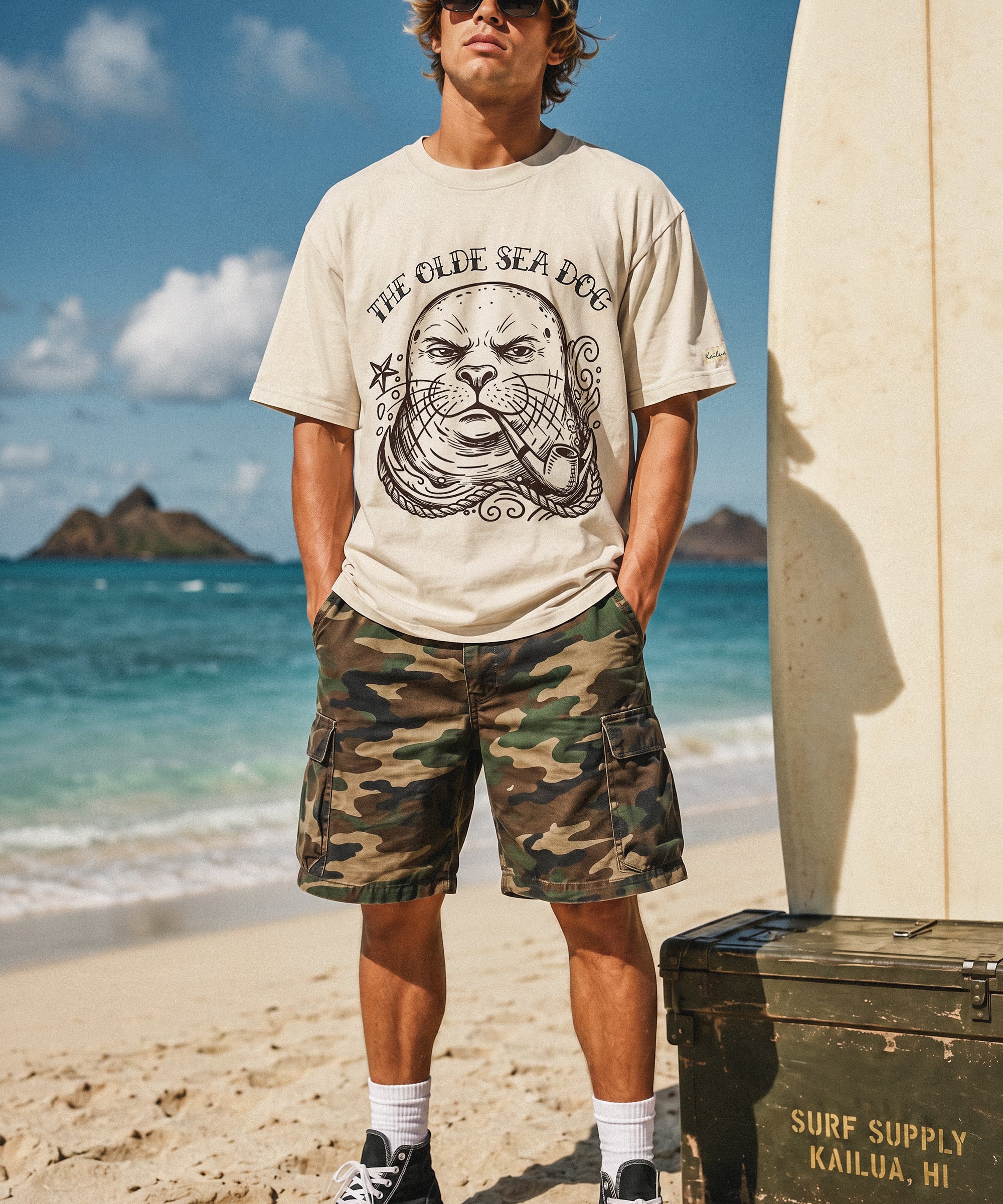 a man standing on the beach next to a surfboard wearing a faded green graphic tee with a monk seal design