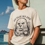 man standing in front of a life guard tower on the beach wearing a faded green graphic tee with a monk seal design