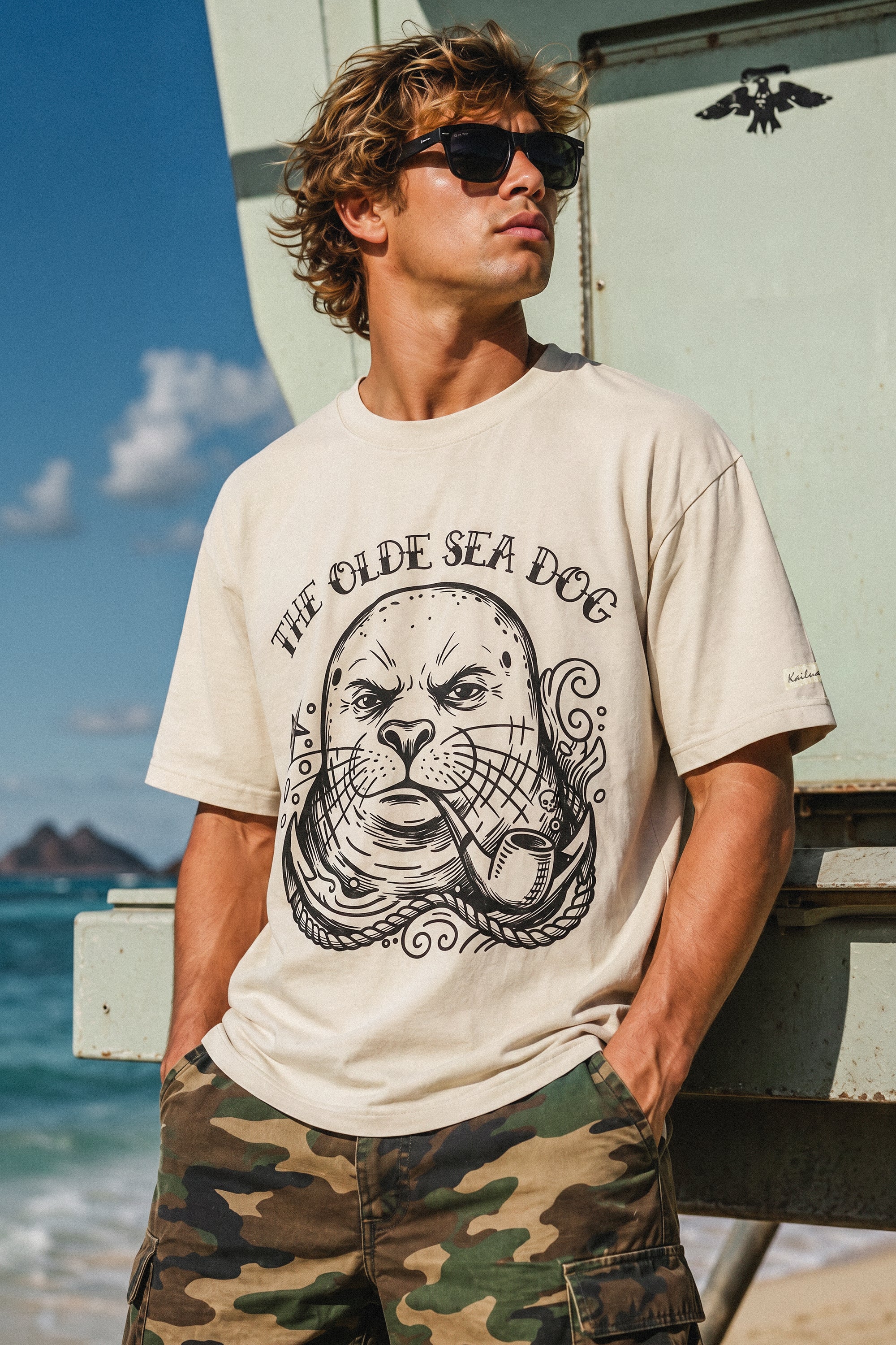 man standing in front of a life guard tower on the beach wearing a faded green graphic tee with a monk seal design