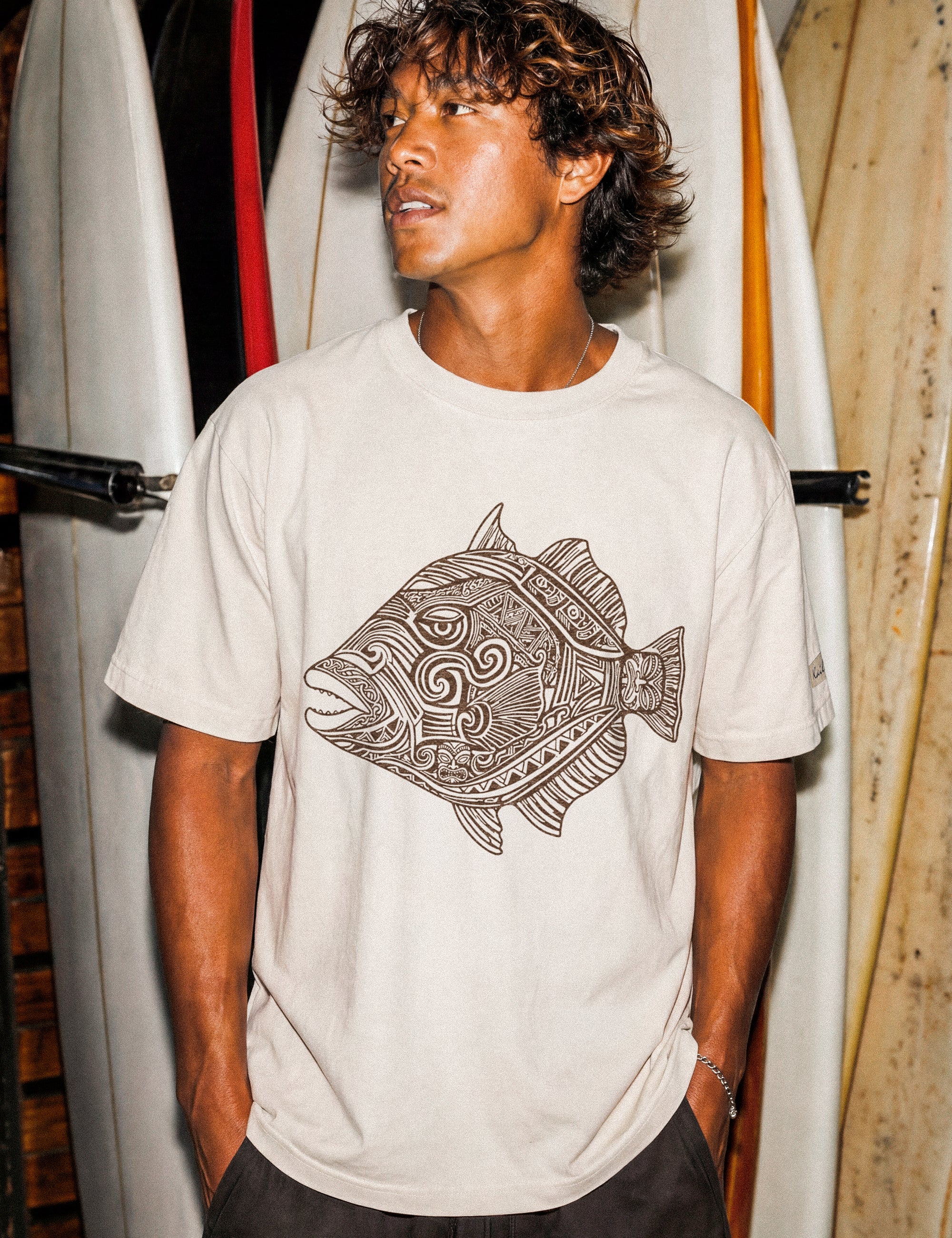a man wearing a pearl colored t-shirt with a fish graphic standing in front of some surfboards