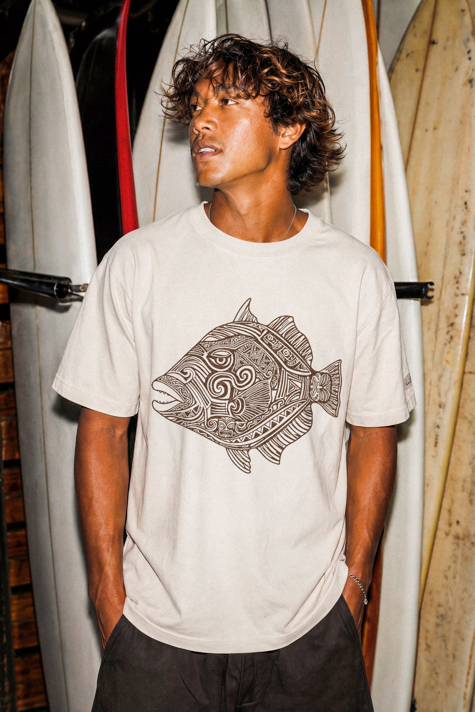 a man wearing a pearl colored t-shirt with a fish graphic standing in front of some surfboards