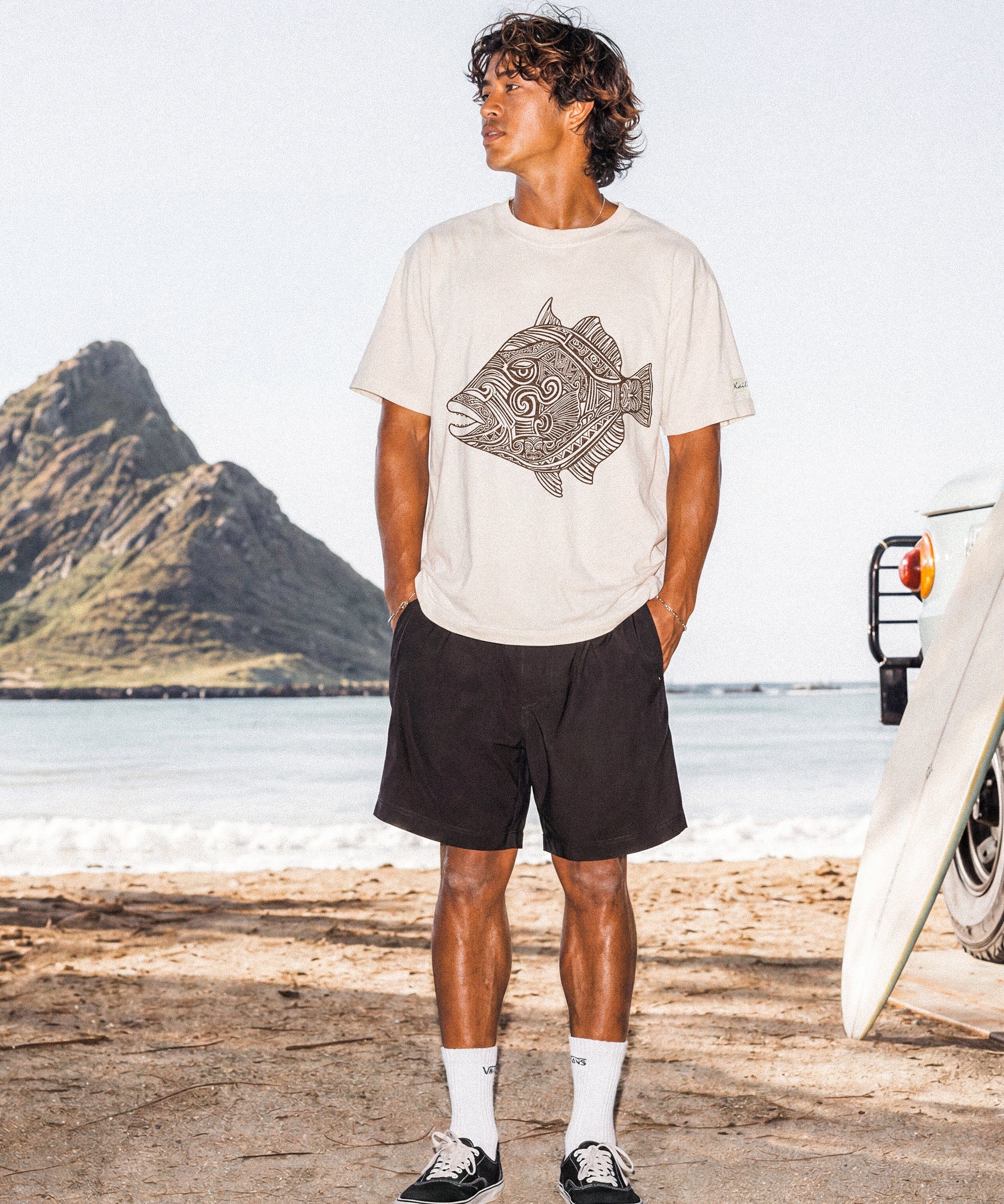 a man wearing a pearl colored t-shirt with a fish graphic standing on a beach