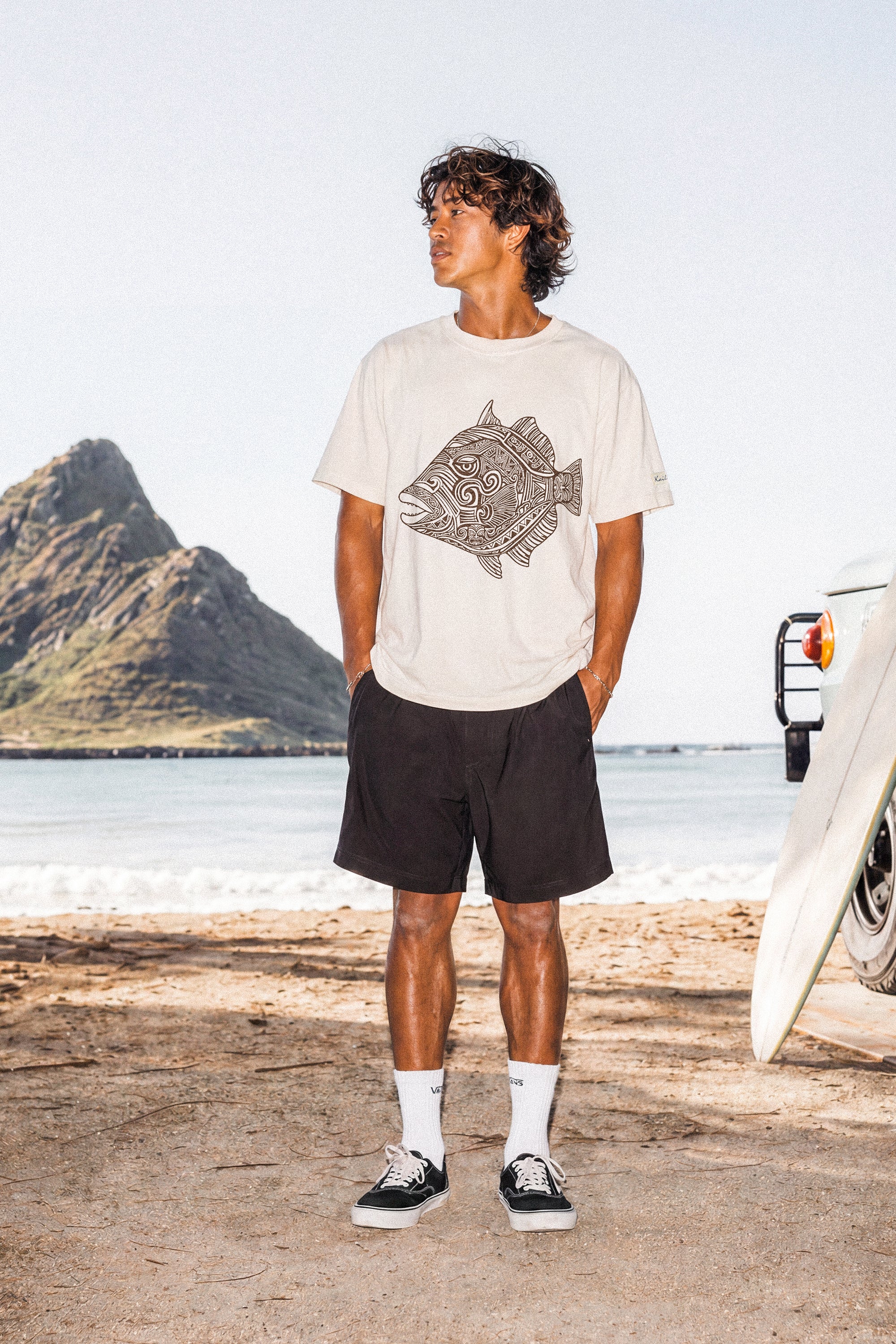 a man wearing a pearl colored t-shirt with a fish graphic standing on a beach