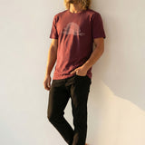 Man wearing a burgundy t-shirt with a white man-o-war with peace signs graphic design against a white wall