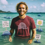 playful beach scene: smiling man stands in shallow water wearing brick-red tee with man-o-war peace graphic, whimsical signs and man-o-war nearby