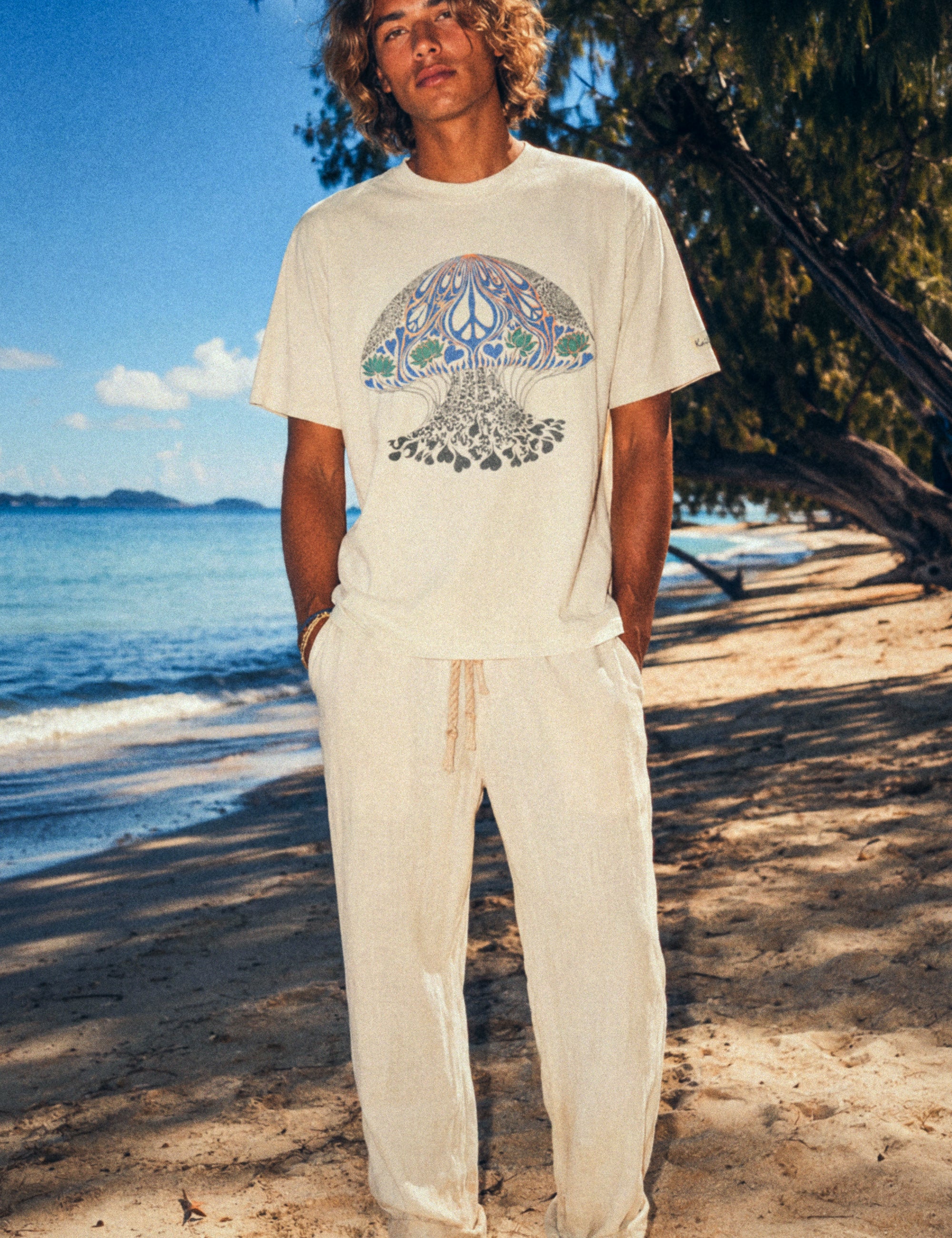 Person standing on a beach wearing a white t-shirt with a colorful design and beige pants.