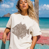 a woman wearing a pearl colored t-shirt with a fish graphic leaning on a wall at the beach