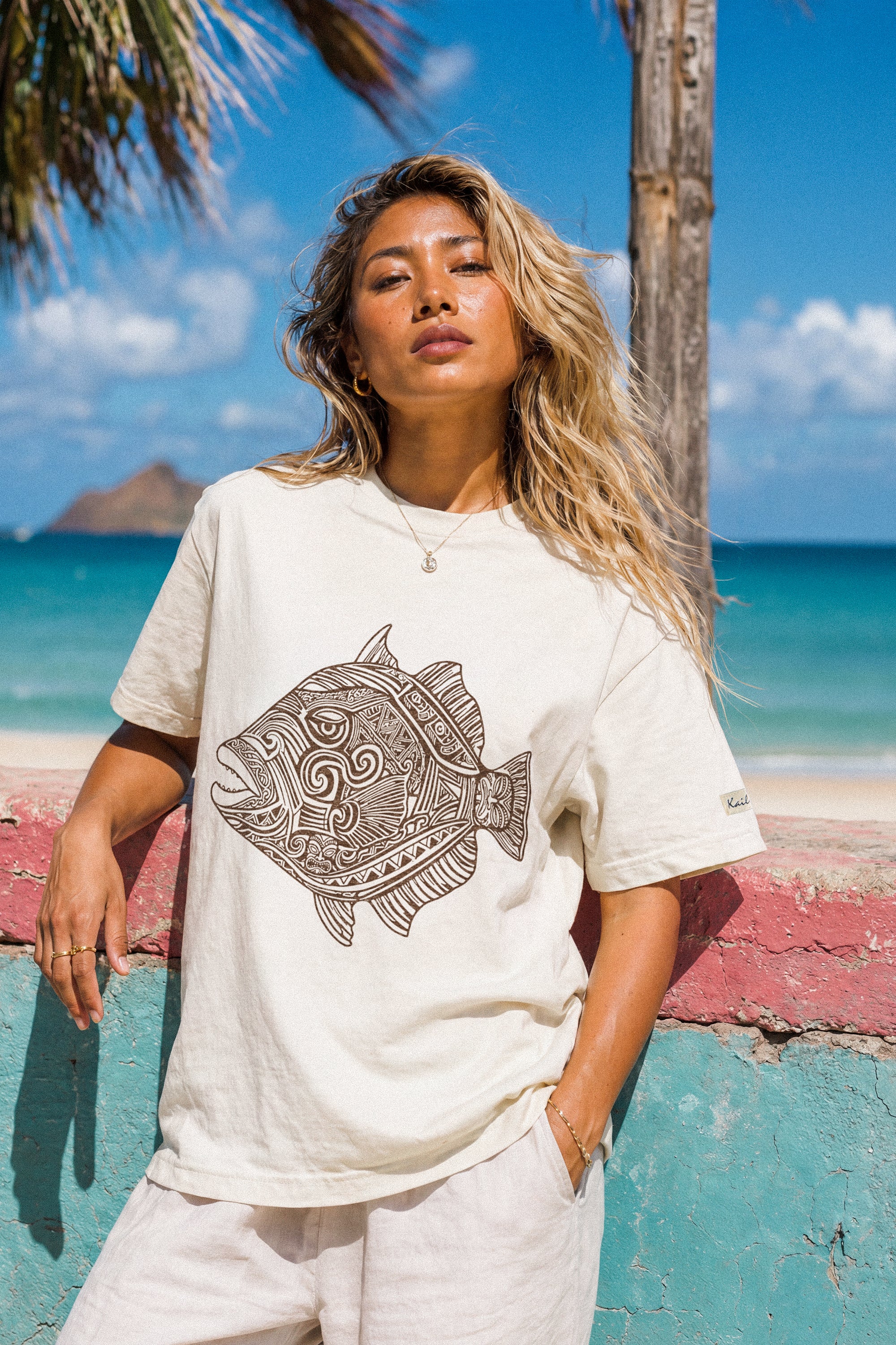 a woman wearing a pearl colored t-shirt with a fish graphic leaning on a wall at the beach