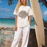 a woman wearing a pearl colored t-shirt with a fish graphic standing on the beach next to a surfboard