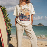 woman standing on the beach next to a surfboard wearing a pearl color t-shirt with a color graphic of a person fishing