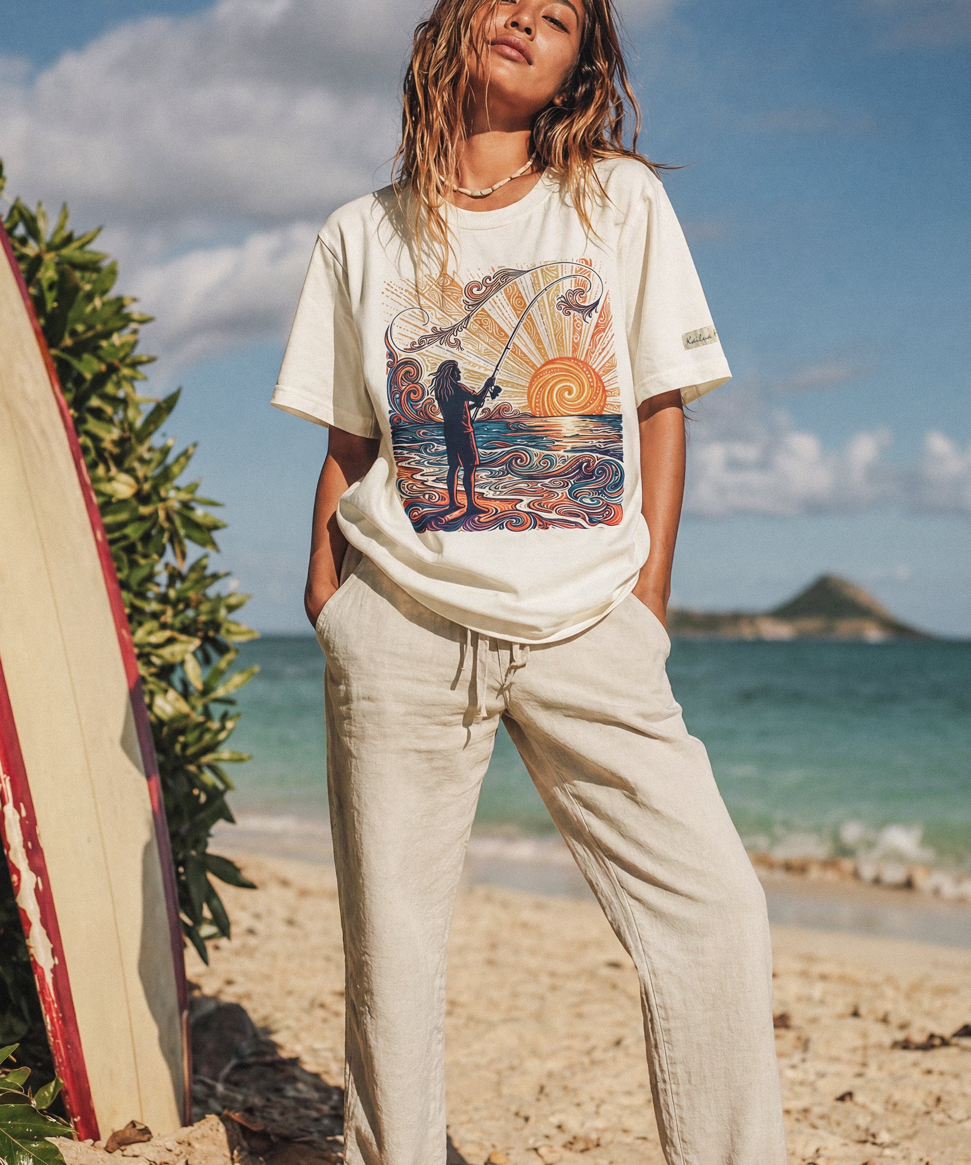 woman standing on the beach next to a surfboard wearing a pearl color t-shirt with a color graphic of a person fishing