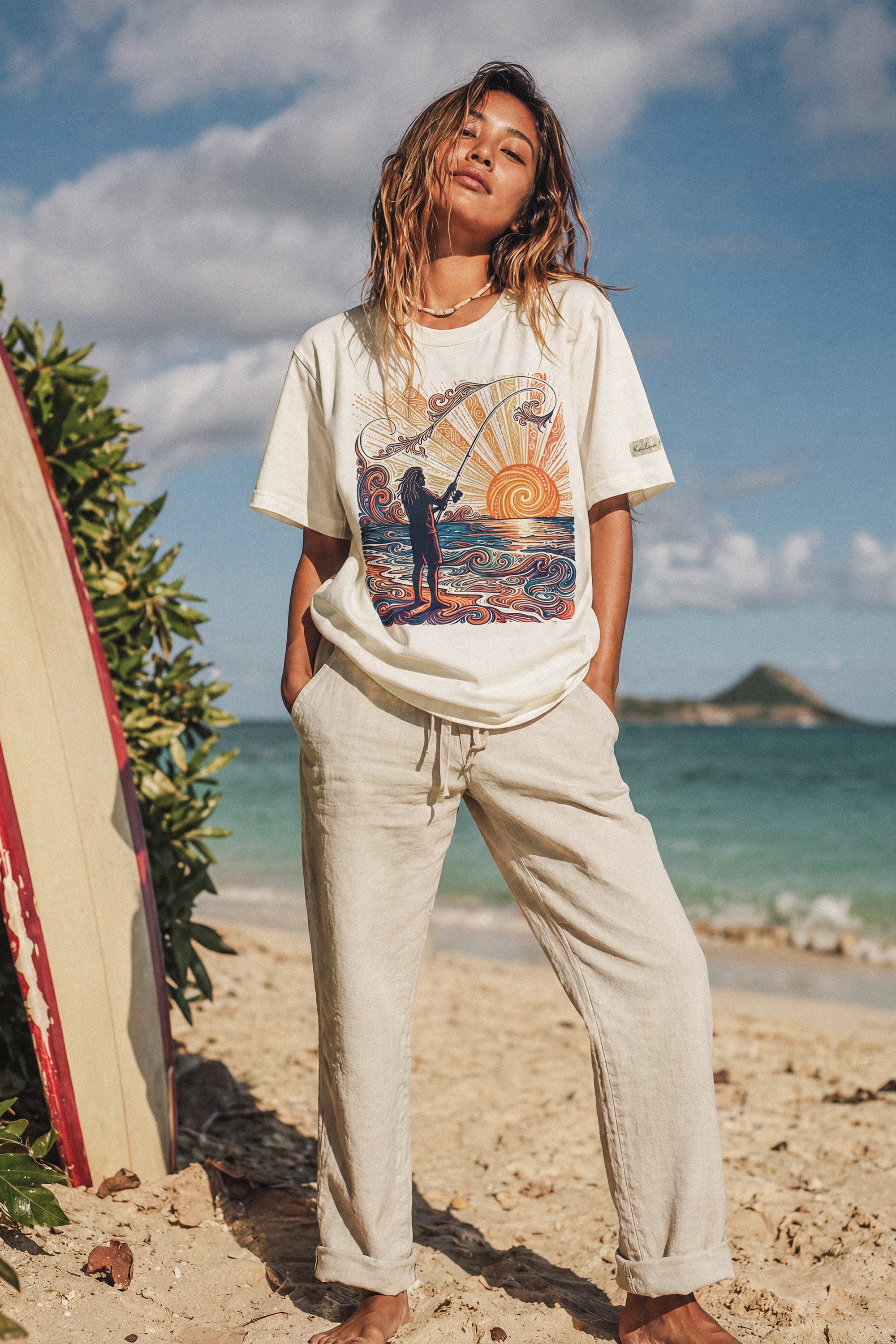 woman standing on the beach next to a surfboard wearing a pearl color t-shirt with a color graphic of a person fishing