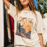a woman standing in front of some surfboards wearing a pearl color t-shirt with a color graphic of a person fishing