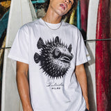 a man wearing a white shirt with a puffer fish graphic standing in front of some surfboards