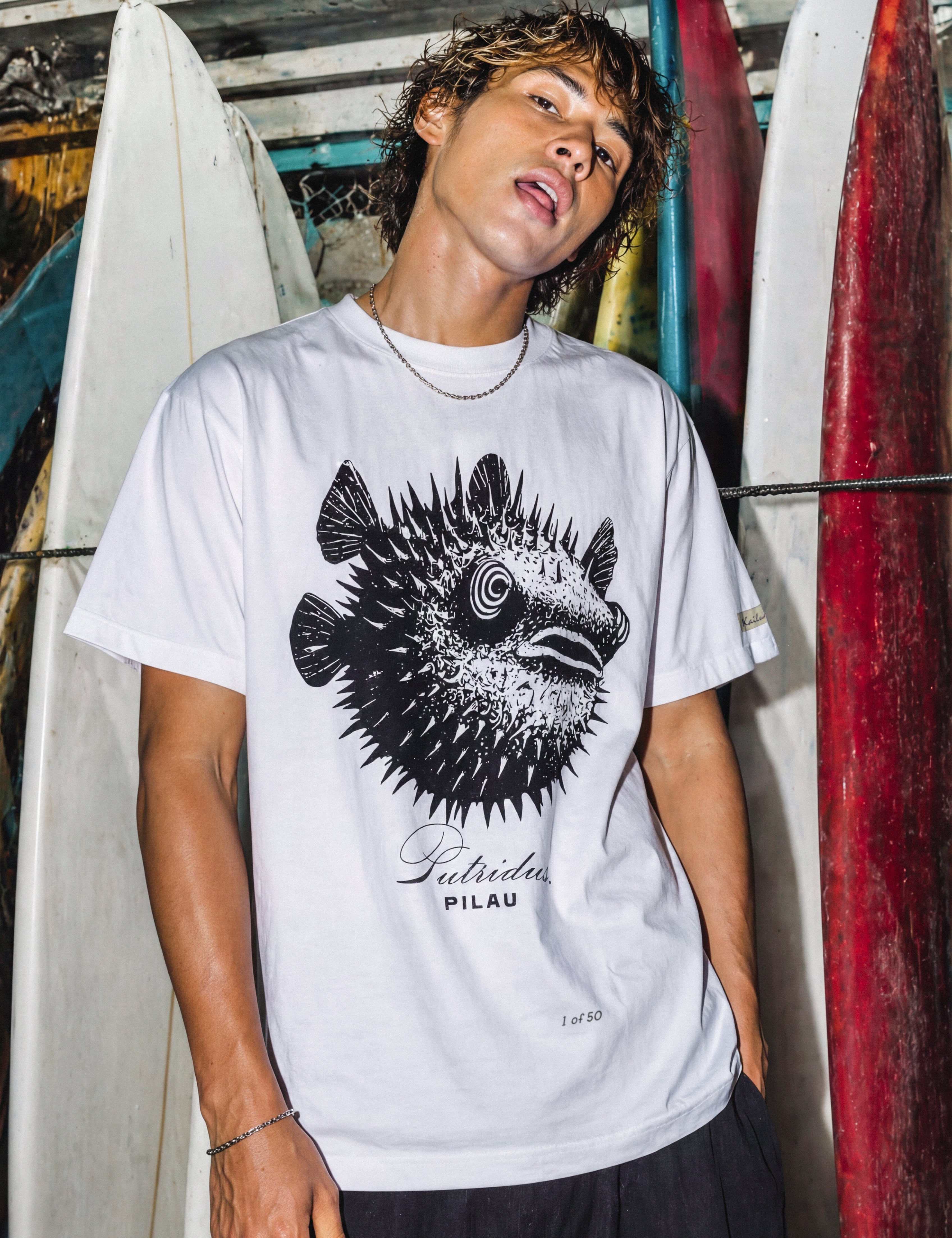 a man wearing a white shirt with a puffer fish graphic standing in front of some surfboards