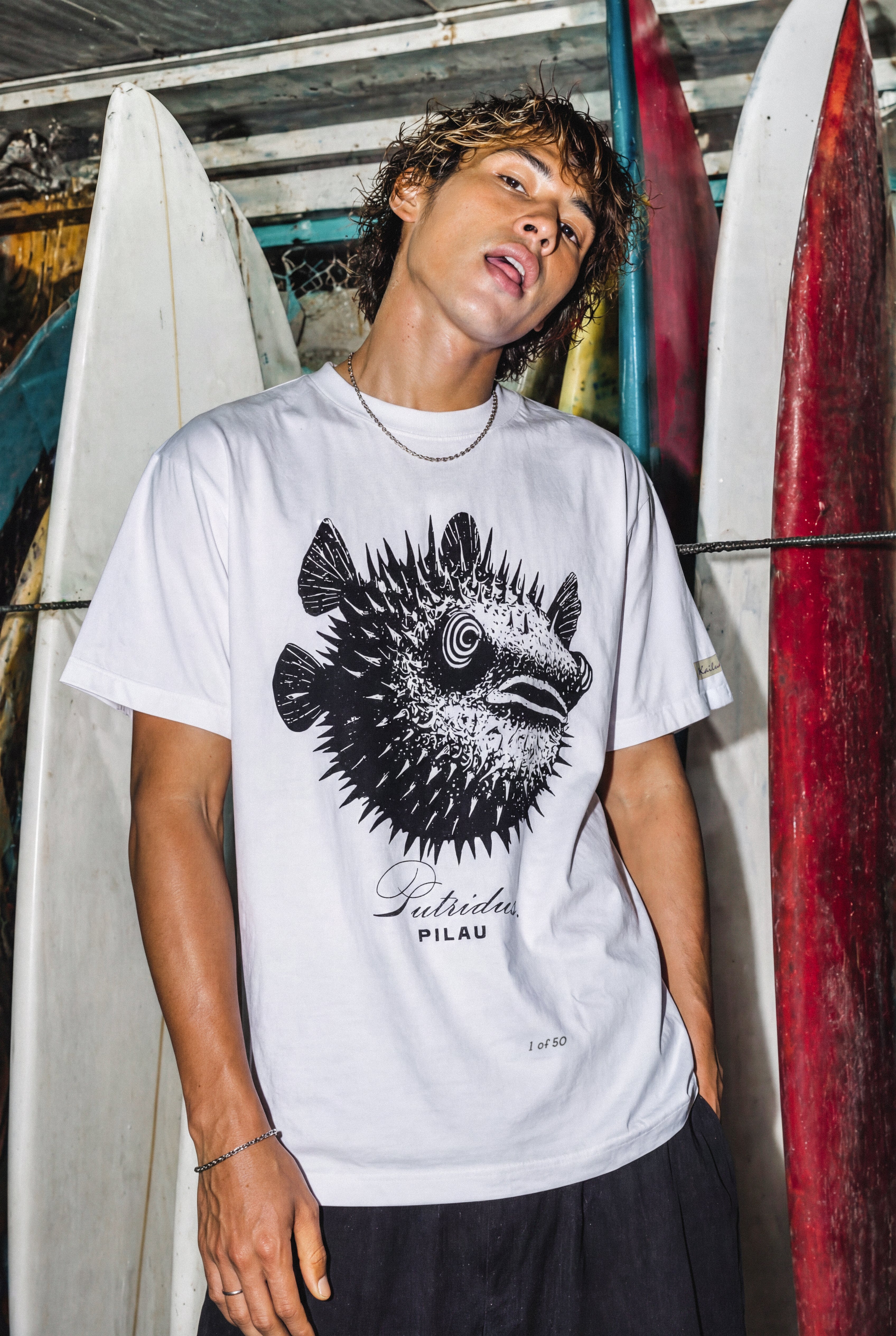 a man wearing a white shirt with a puffer fish graphic standing in front of some surfboards