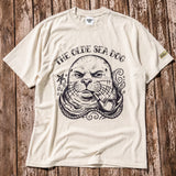 a faded green graphic tee with a monk seal design laying on a wood floor