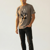 Man wearing a brown graphic t-shirt with a design of a man giving the shaka hand sign on a plain background