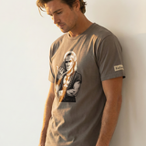 Man wearing a brown t-shirt with a graphic design of a man giving the shaka hand sign against a white wall